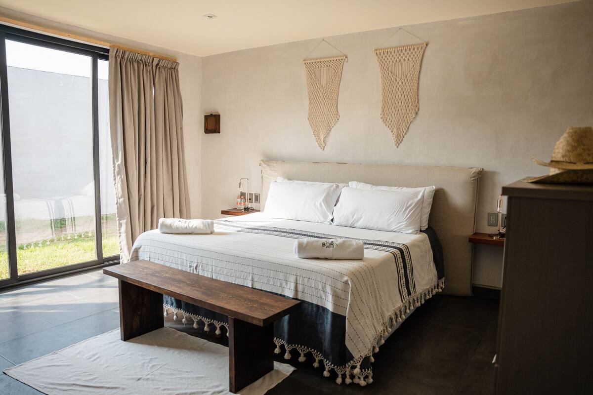 A modern bedroom with a large bed, a bench, a bedside table, and a view of the outdoors.