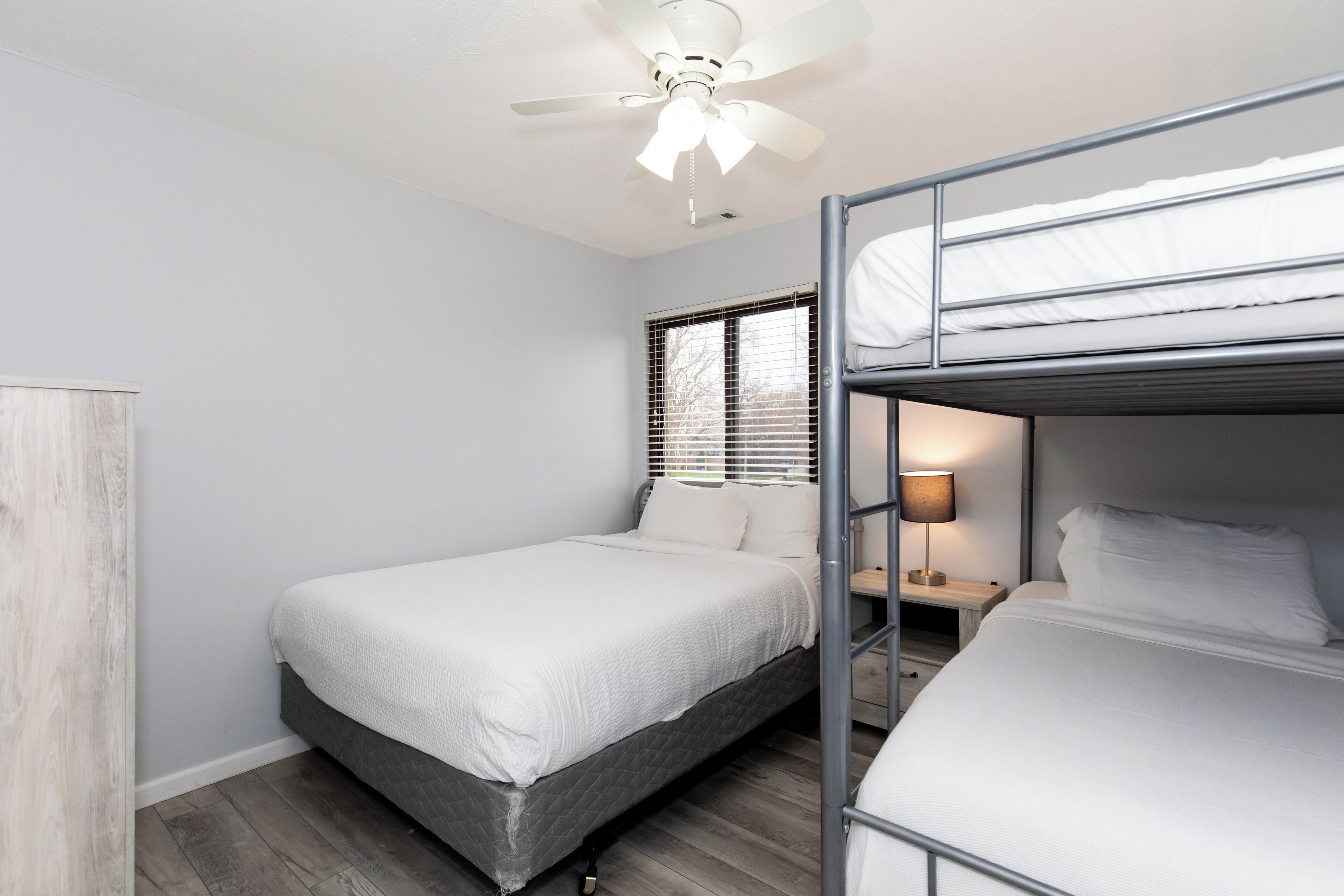 A bedroom with a single bed, a bunk bed, a ceiling fan, a window with blinds, and a nightstand with a lamp.