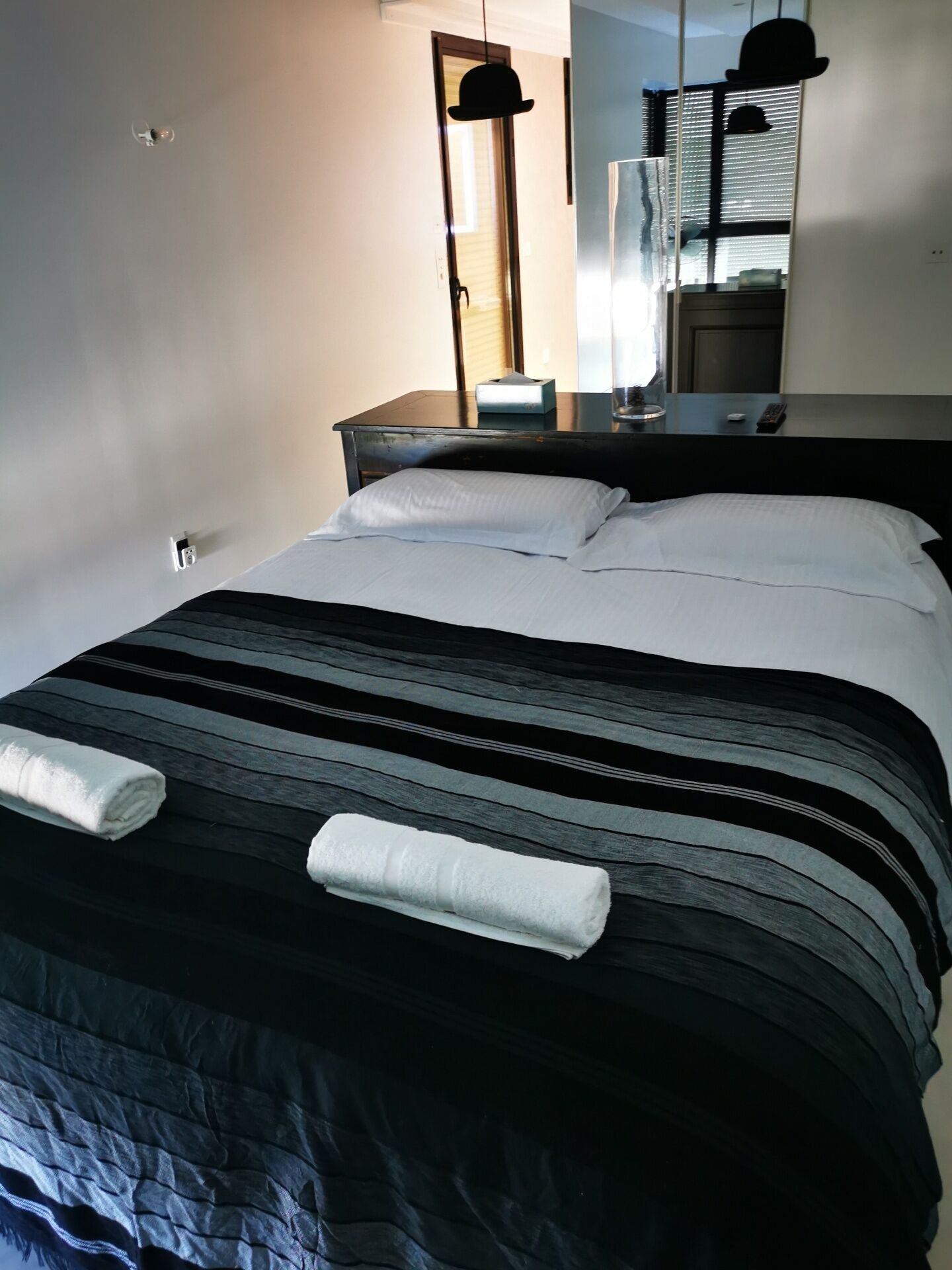 Un lit bien fait, recouvert d’une couverture à rayures et agrémenté de deux serviettes enroulées, une table de chevet avec une lampe et un miroir qui reflète une fenêtre avec des stores.
