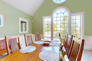 A dining room with a wooden table set for six, surrounded by chairs, large windows with a view of trees, and a small window on the left. - Stunning lakefront property with beach access, a dock, spacious yard, & W/D (Moultonborough)