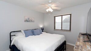 A bedroom with a bed, a ceiling fan, a window with blinds, and a desk with a lamp.