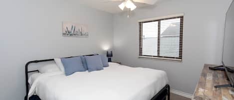 A bedroom with a bed, a ceiling fan, a window with blinds, and a desk with a lamp.