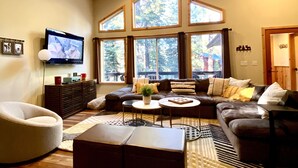 A spacious living room with a large sectional sofa, a round coffee table, and a comfortable armchair. There are multiple windows providing natural light and a view of the outdoors. - Cozy Tahoe Donner Retreat – Hot Tub, Fireplace & Near Ski Resorts! (Truckee)