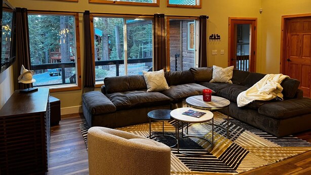 A cozy living room with a sectional sofa, a coffee table, and a patterned rug. Large windows offer a view of the outdoors. - Cozy Tahoe Donner Retreat – Hot Tub, Fireplace & Near Ski Resorts! (Truckee)