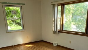 Una habitación con piso de madera, dos ventanas con cortinas y vista a áreas verdes.
