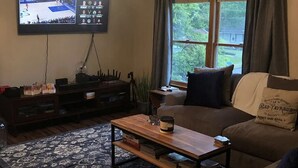Una sala de estar con televisor mostrando un partido deportivo, sofá, mesa de centro y ventana con cortinas.