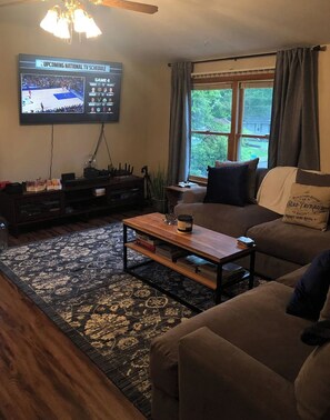 Una sala de estar con televisor mostrando un partido deportivo, sofá, mesa de centro y ventana con cortinas.