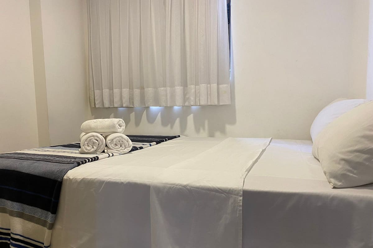 A neatly made bed with white bedding, a striped blanket, and rolled towels on the side.