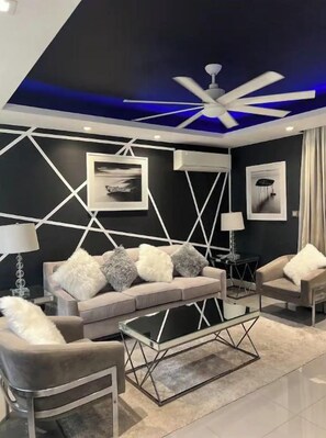 Un salon moderne avec un motif géométrique sur le mur, un ventilateur de plafond et une table basse en verre.