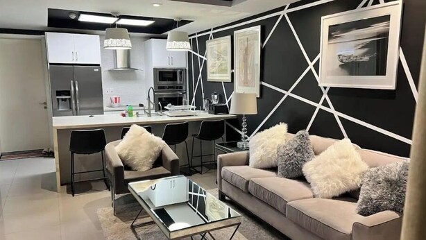 A modern living room with a sofa, coffee table, and chairs. A kitchen with a dining area is visible in the background. - Luxurious 1 bedroom condo. On the South (Kingston)