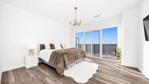 A bedroom with a large bed, a nightstand, a white rug, and a view of the sky through large windows.