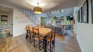 A spacious dining area with a wooden table and chairs, a staircase leading to another level, and a living area with a sofa and television. - Secluded Retreat w Hot Tub, FirePit & Pool Access (Hedgesville)