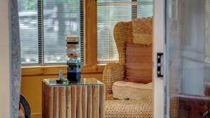 A room with a wooden side table holding a stack of books, a woven chair, and a window with blinds. - Secluded Retreat w Hot Tub, FirePit & Pool Access (Hedgesville)