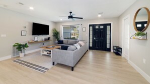 A modern living room with a grey sofa, a flat-screen TV, a ceiling fan, and a large window. - ☆New☆6min to 3 Sisters Springs☆Spacious, Fun with Game room (Crystal River)