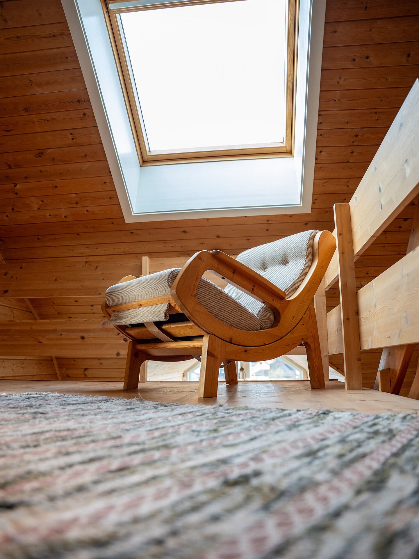 A room with wooden walls, a sloped ceiling, and a skylight. There is a woven chair and a wooden table.