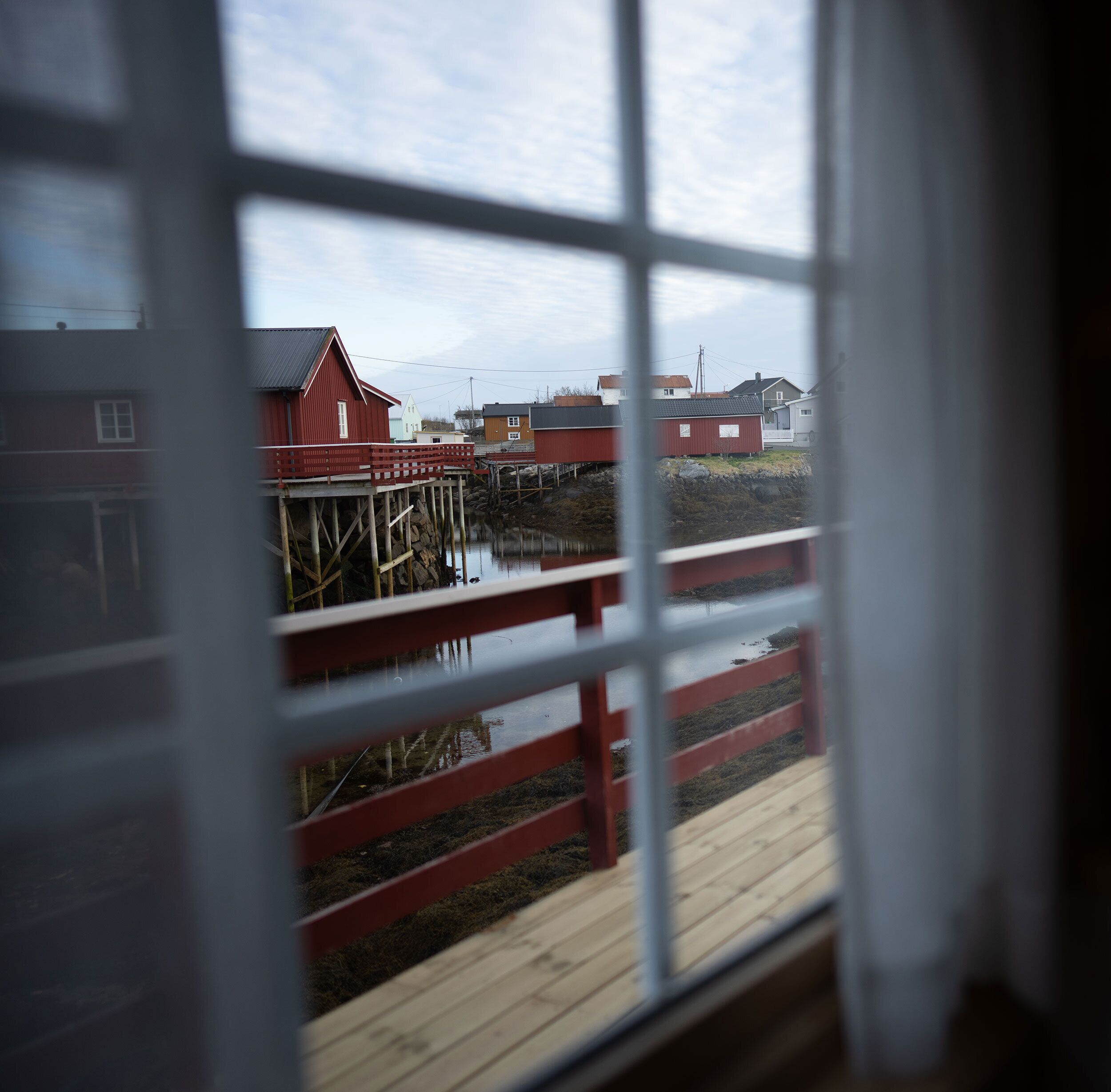 Une vue à travers une fenêtre donnant sur une terrasse en bois et un bâtiment rouge avec une fenêtre blanche.