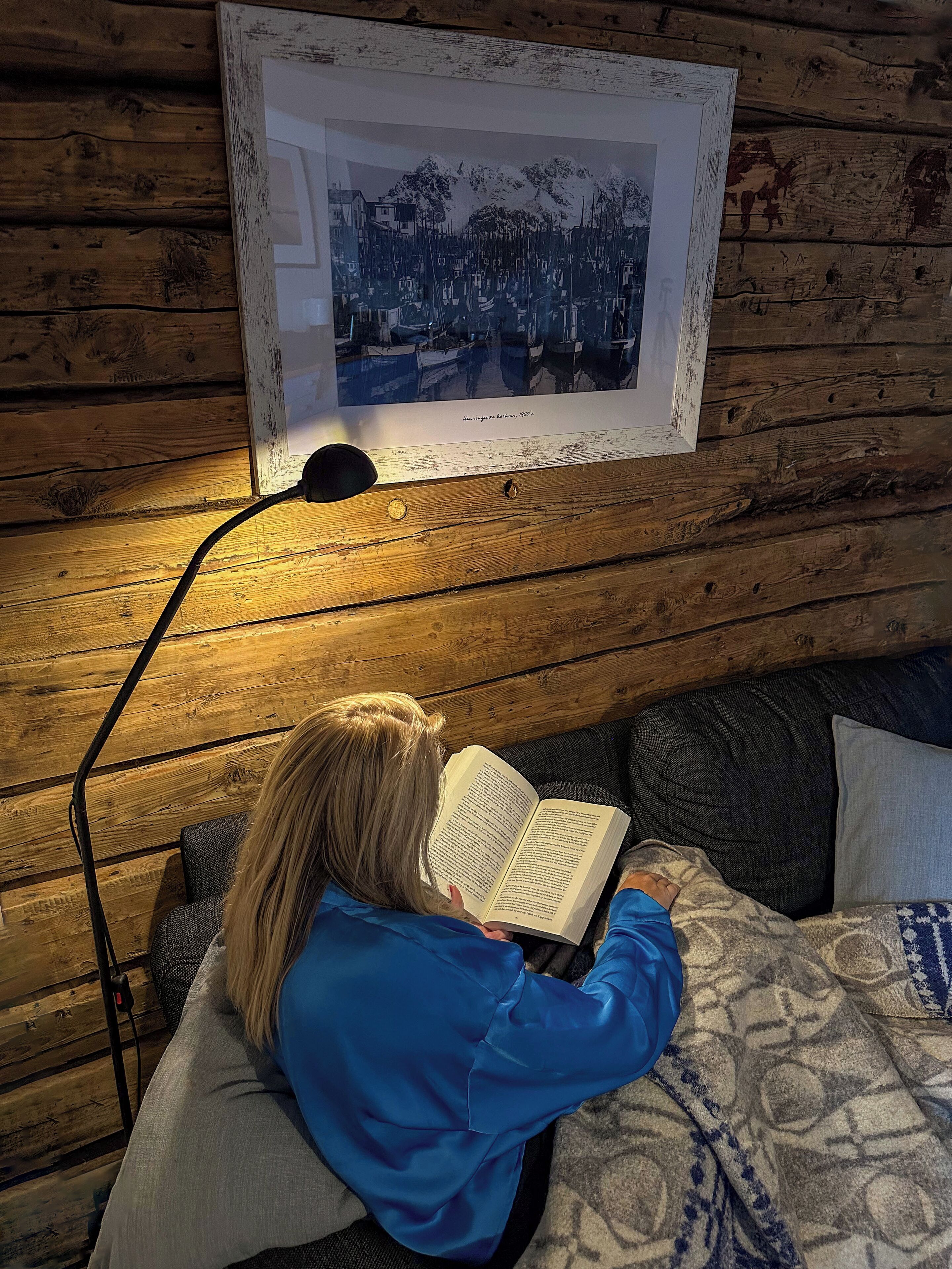 Eine Person liest ein Buch in einem Zimmer mit Holzwänden, einem gerahmten Bild an der Wand und einer Lampe auf einer Holzfläche.