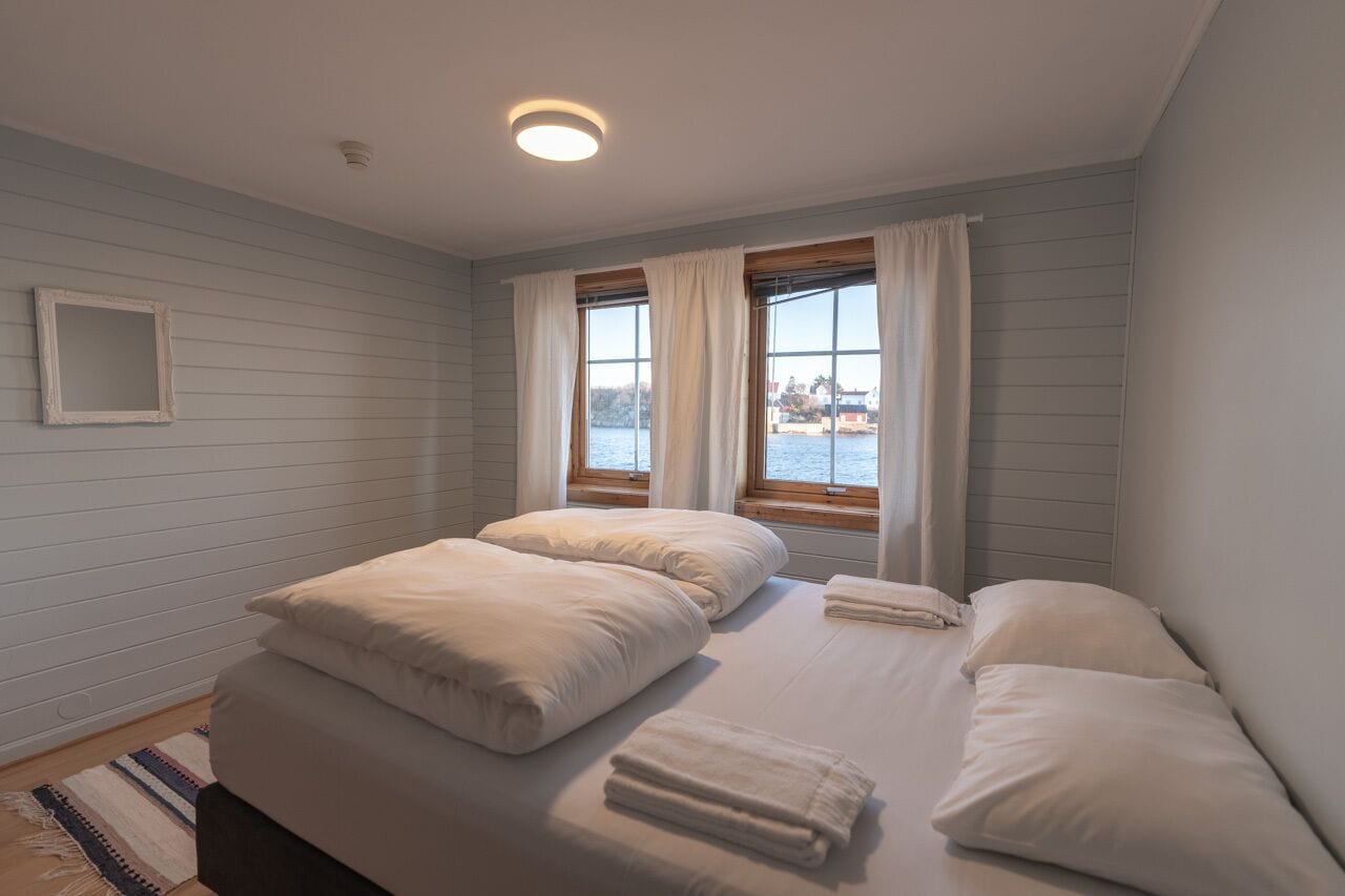 A bedroom with two beds, a window with curtains, a small mirror on the wall, and a view of the water outside.