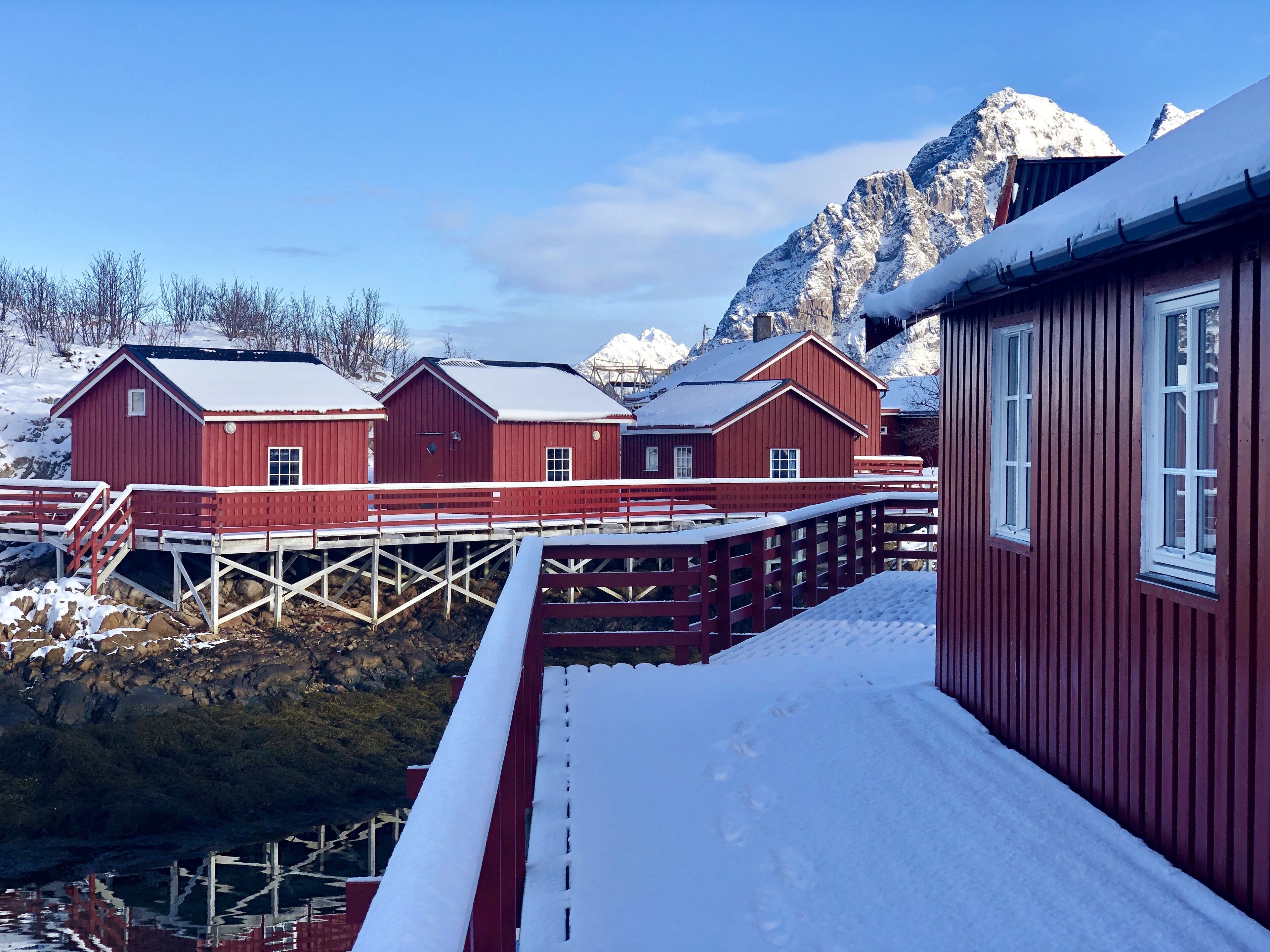 A red wooden building with a snow-covered roof, situated near a body of water with a wooden dock.