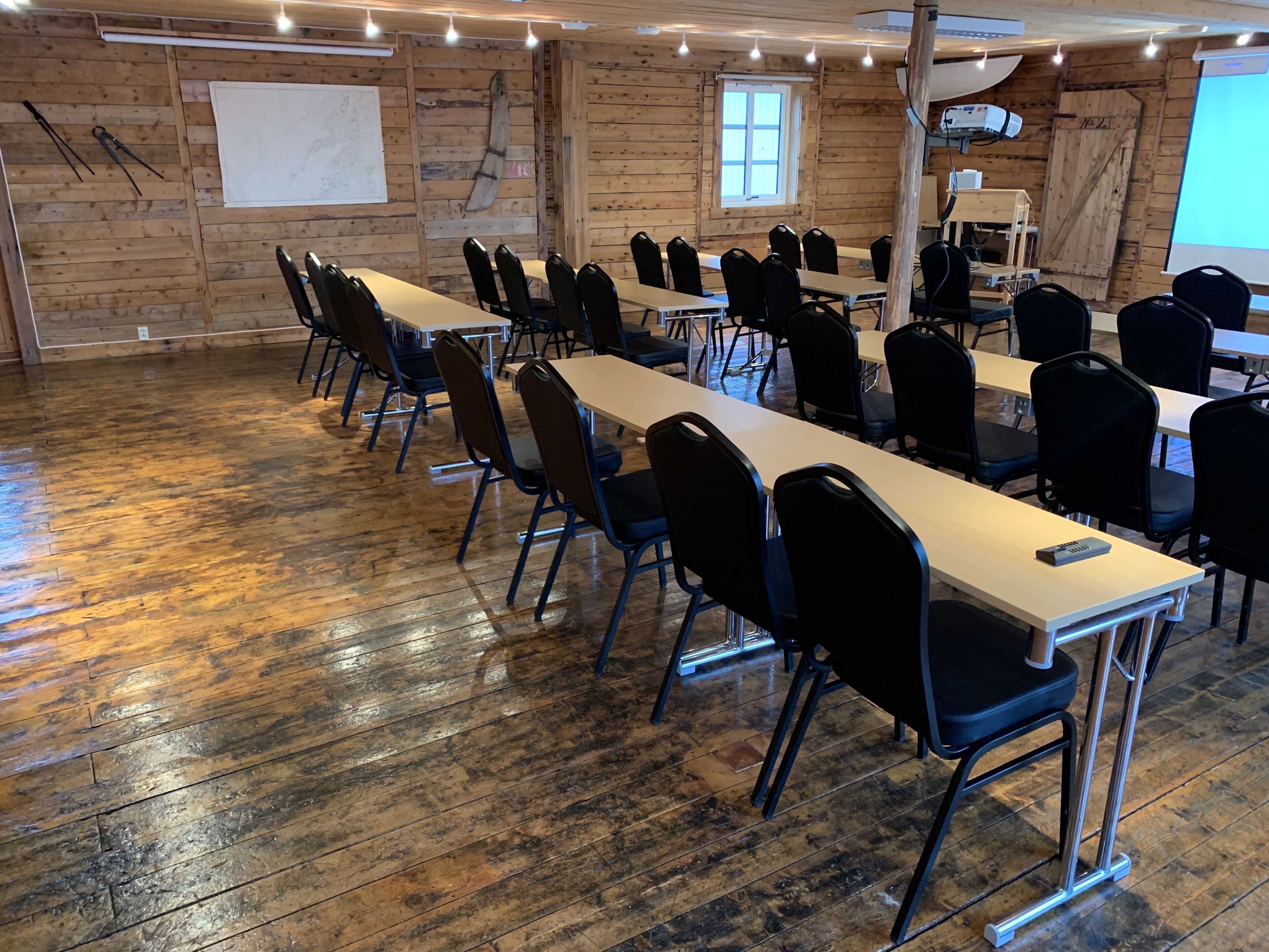 A conference room with long tables and chairs, wooden walls, and a projector screen.
