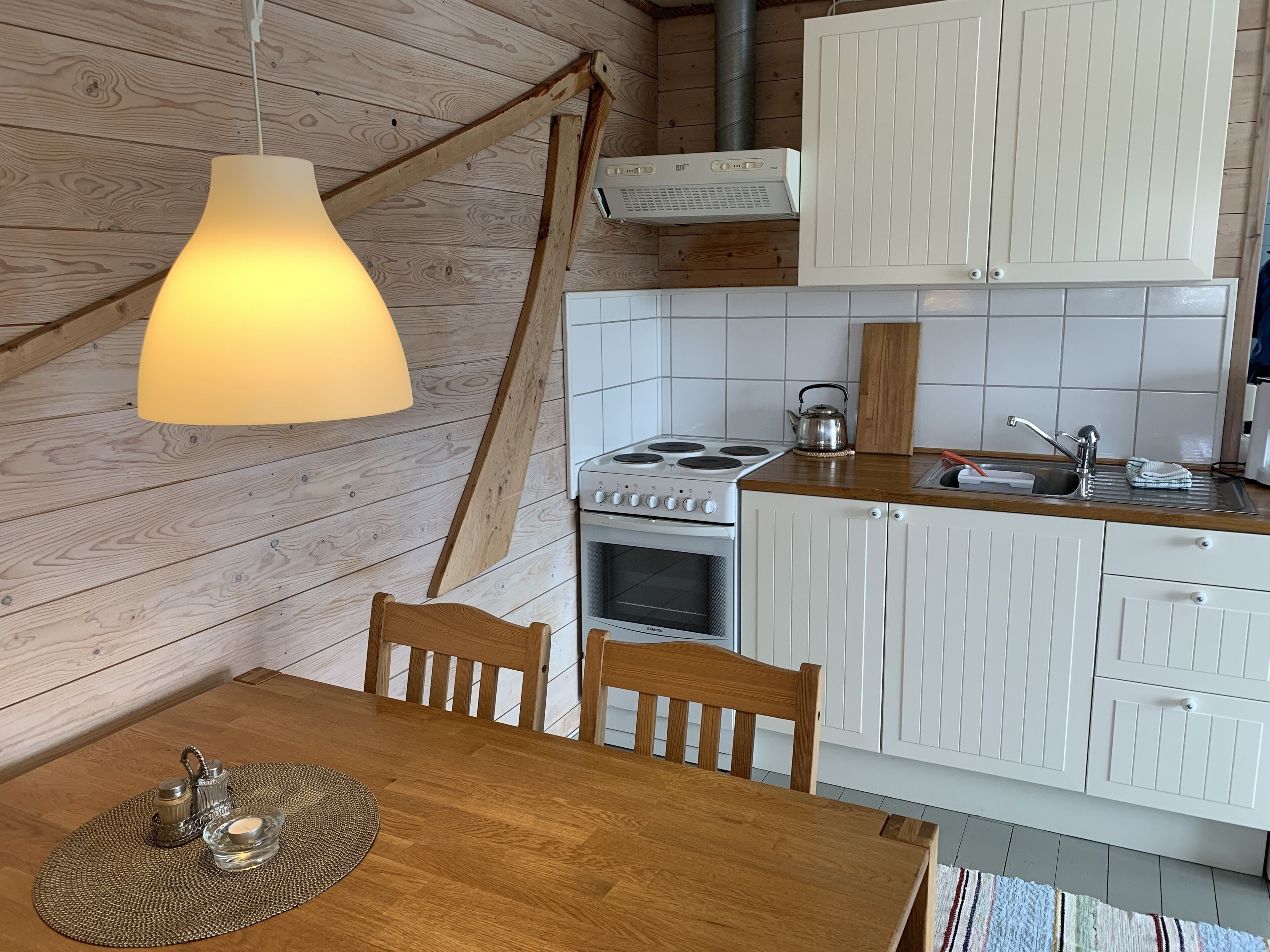 Une cuisine avec des murs en bois, des armoires blanches, un fourneau et un luminaire suspendu.