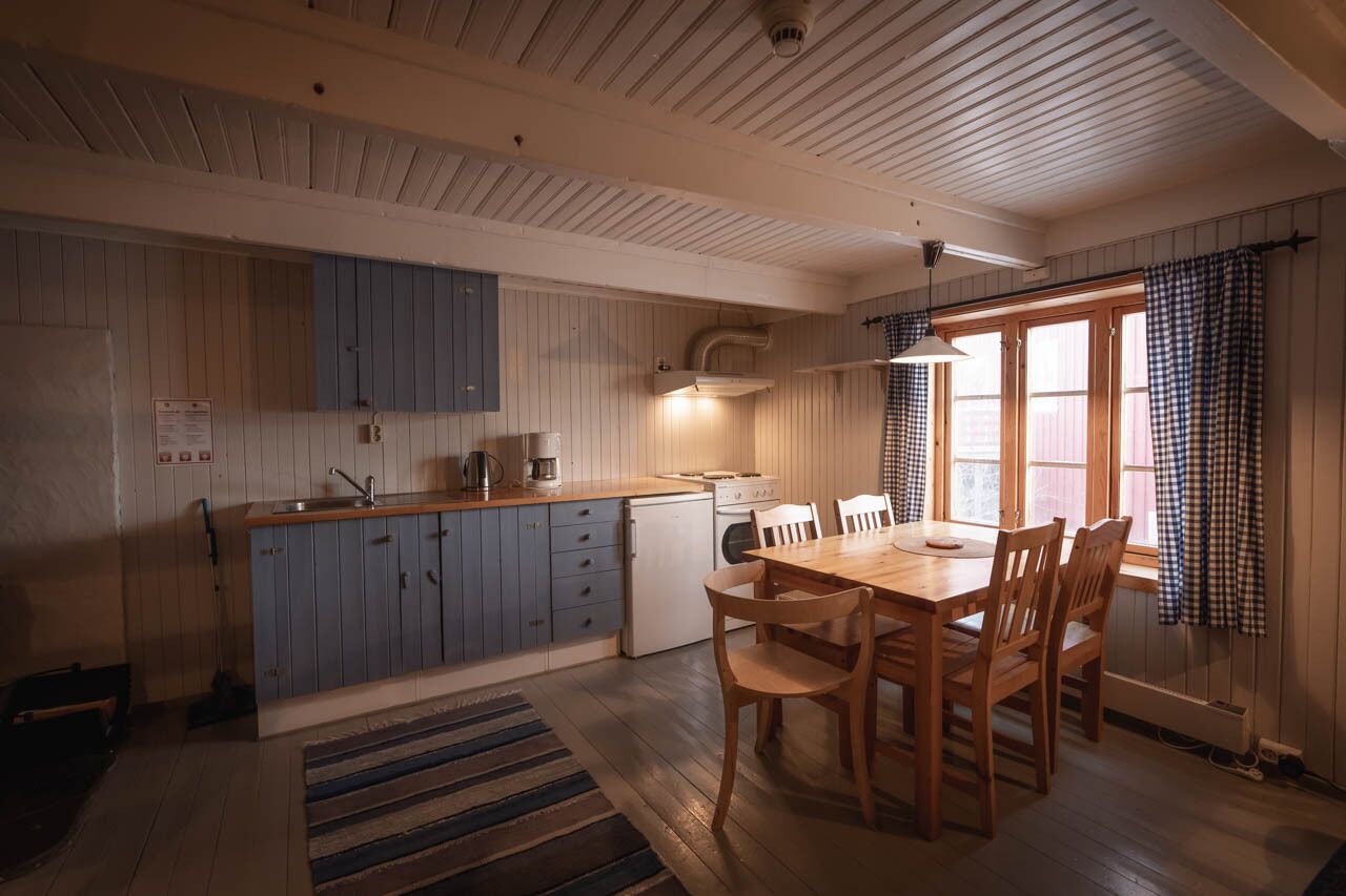 Une cuisine de taille réduite, dotée de meubles en bois, d’une table à manger et d’une fenêtre ornée de rideaux à carreaux.