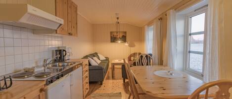 A compact kitchen with wooden cabinets, a dining table, and a view of the sea through a large window.