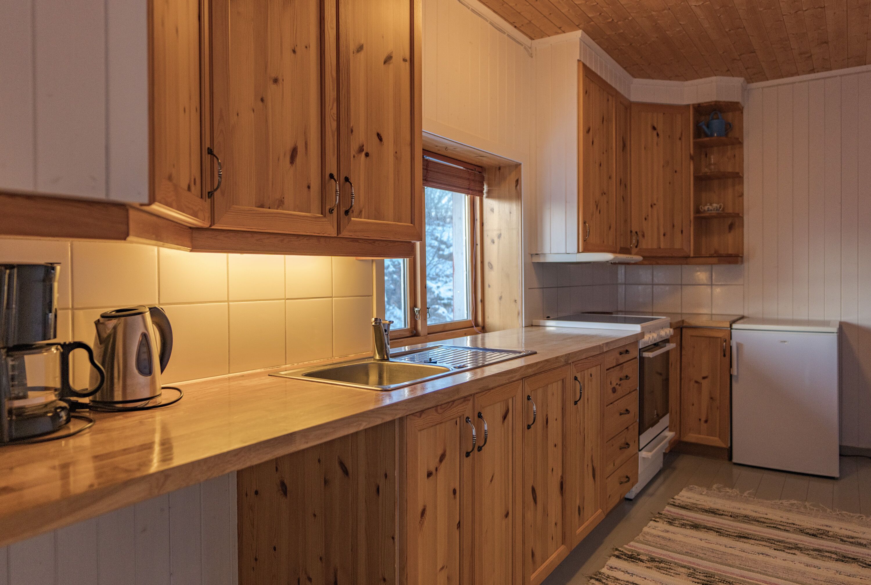Una cocina con armarios de madera, un lavamanos de acero inoxidable, una tetera y un refrigerador.