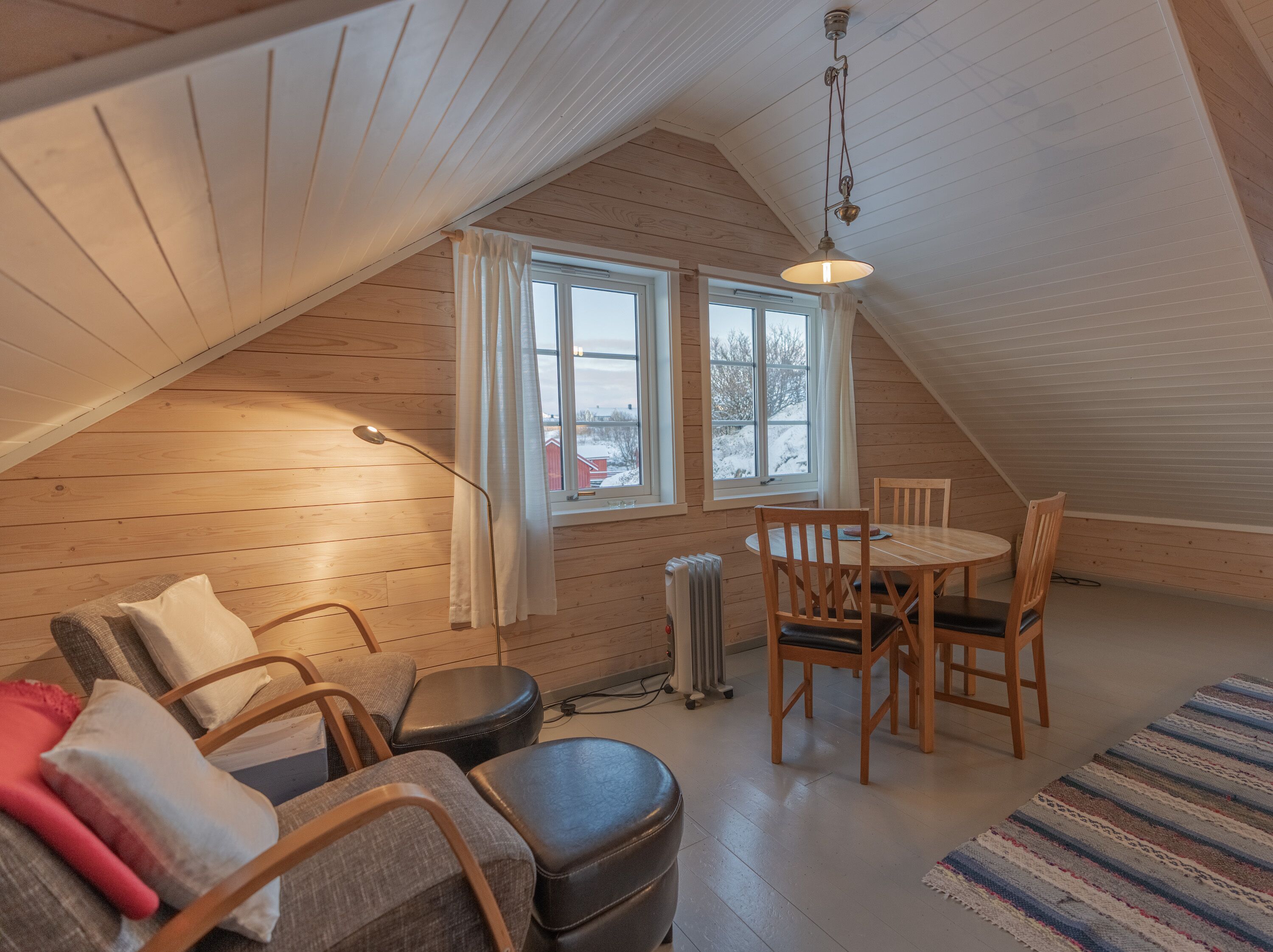 Une pièce mansardée chaleureuse avec un plafond en pente en bois, une table à manger avec des chaises, un lampadaire et une vue sur un paysage enneigé par deux fenêtres.