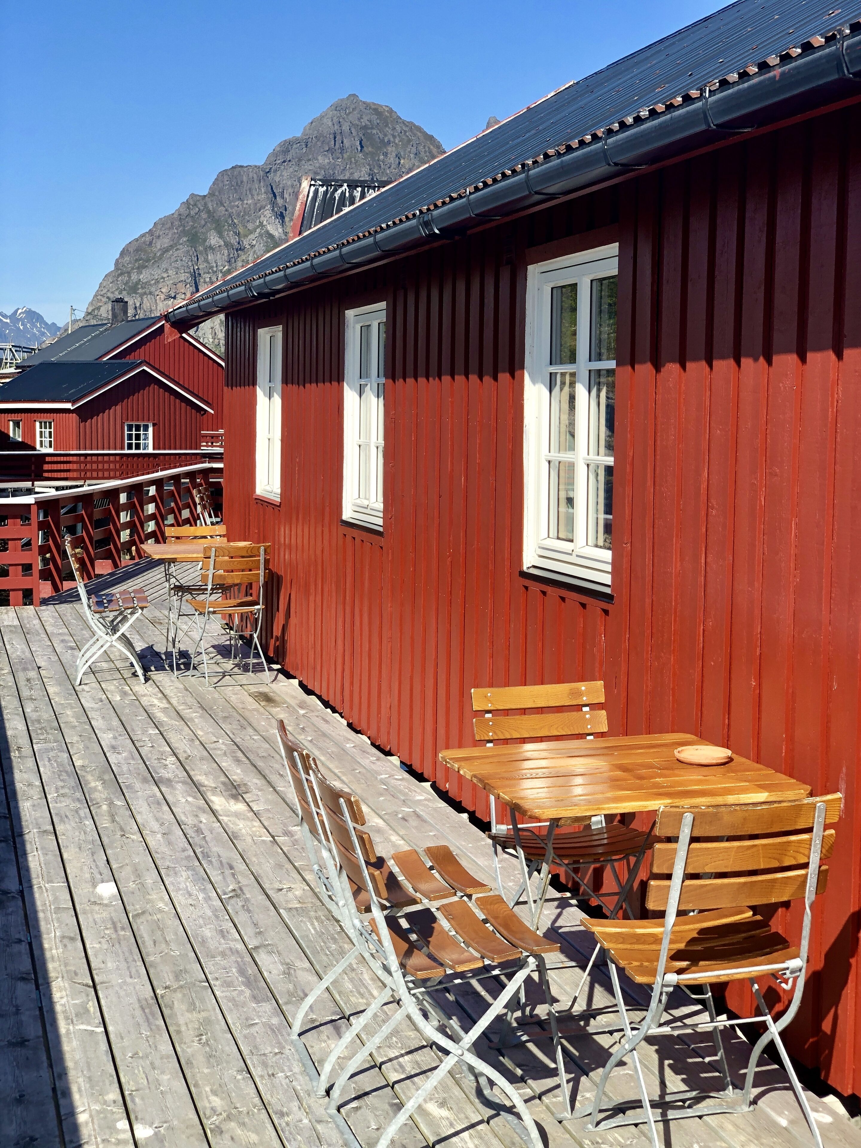 A row of red wooden buildings with white windows, wooden tables, and metal chairs on a wooden deck.