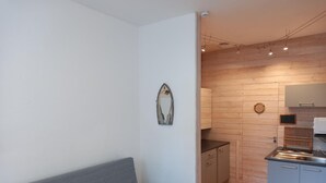 Un espace de vie compact comprenant un canapé gris, une petite table ronde, une kitchenette avec des panneaux en bois et un miroir mural.