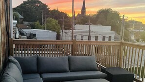 A wooden deck with a gray sectional sofa and a small table, overlooking a cityscape with a church spire during sunset.