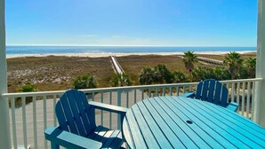 A balcony with a view of the beach, two blue Adirondack chairs, and a blue table.