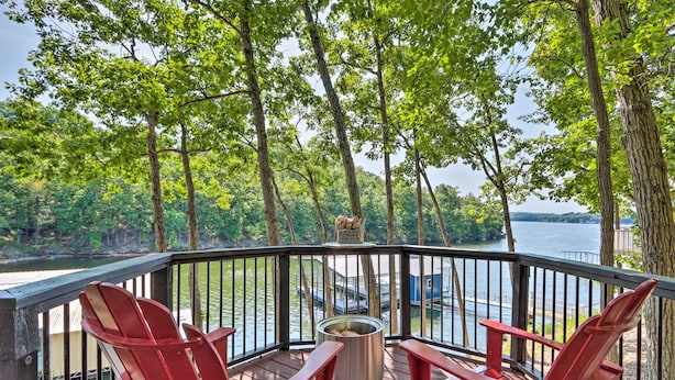 Une terrasse avec des fauteuils Adirondack rouges, un foyer en métal et une vue sur un lac où naviguent des bateaux.