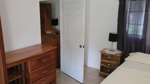 Quarto com cômoda de madeira, cama com colcha branca, mesa de cabeceira com abajur e porta que dá para outro espaço.