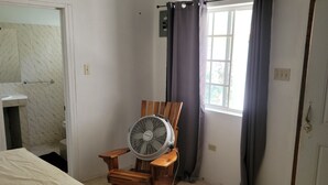 Quarto com uma cama, uma cadeira de madeira com ventilador, uma janela com cortinas e uma porta.