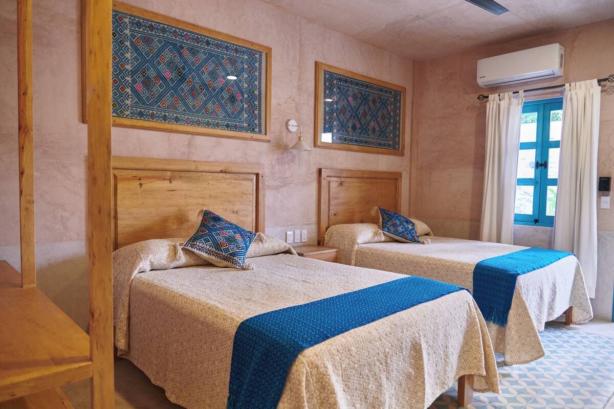 Une chambre avec deux lits, chacun doté d’une tête de lit en bois et d’une couvre-lit bleue. On y trouve deux climatiseurs fixés au mur et une fenêtre avec des rideaux.