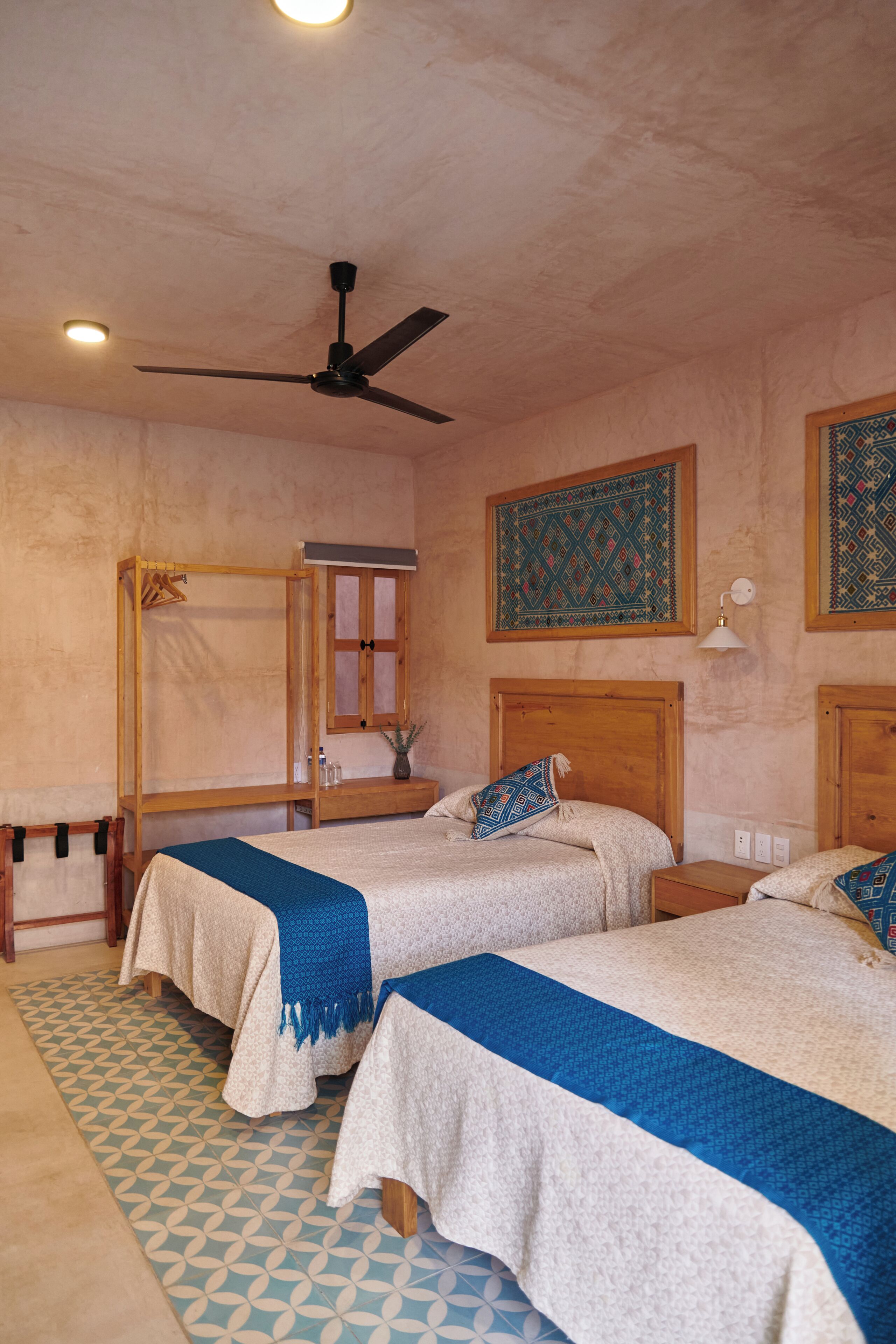 A room with two beds, a ceiling fan, a wooden wardrobe, and patterned wall hangings.