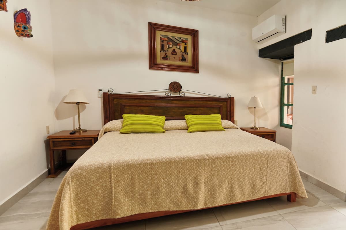 Un lit bien fait, recouvert d’une couvre-lit à motifs et agrémenté de deux oreillers, avec deux tables de chevet surmontées de lampes, dans une pièce où un tableau encadré orne le mur et où un climatiseur est fixé au mur.
