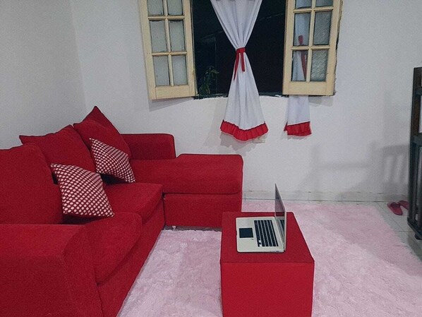 A red sofa with patterned pillows, a laptop on a matching ottoman, and a window with curtains.
