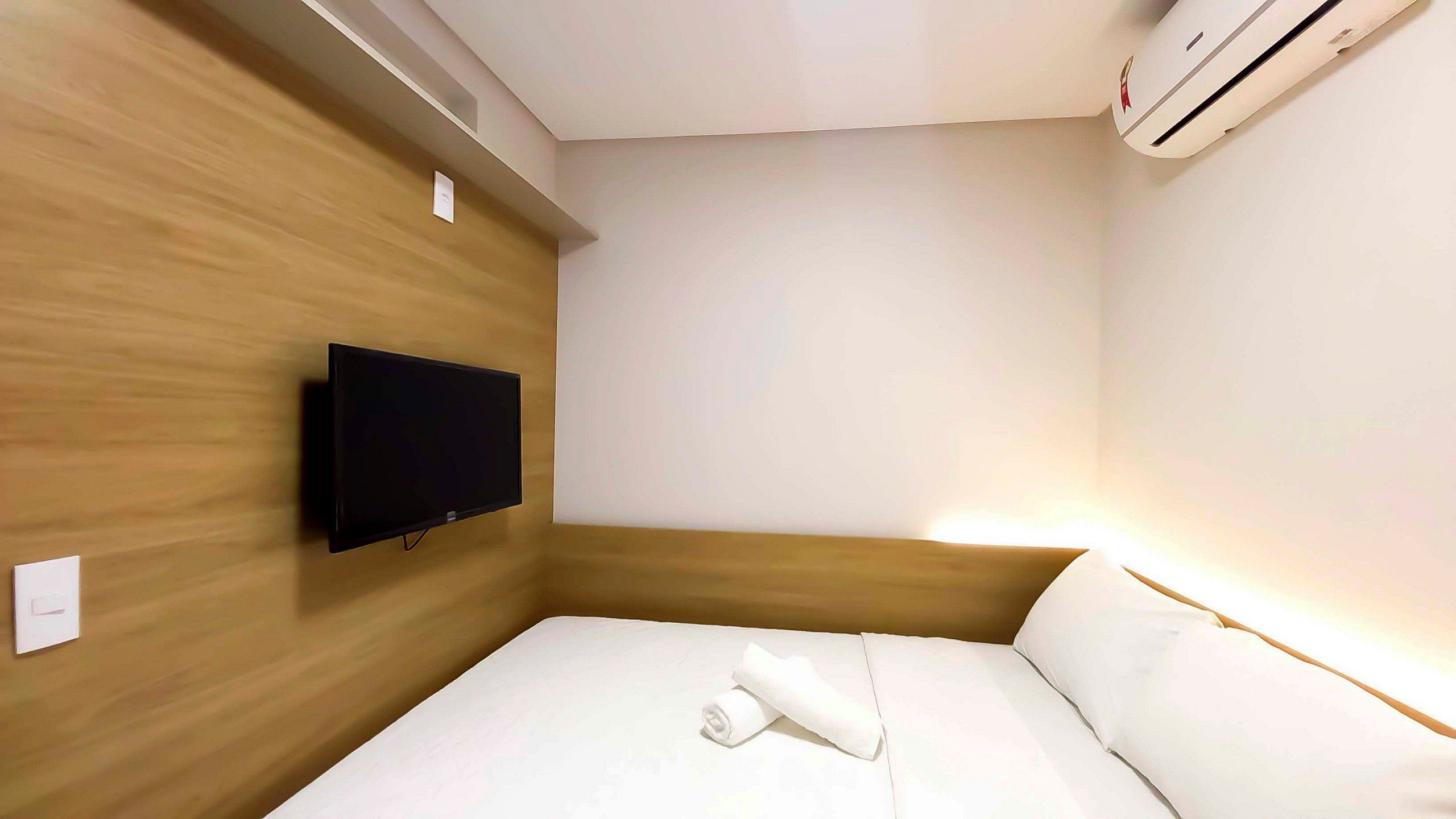 A hotel room with a bed, a flat-screen TV mounted on the wall, and an air conditioning unit.