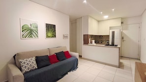 Een woonkamer met een bank, een keukenhoek met een koelkast, en een deur naar een andere kamer.