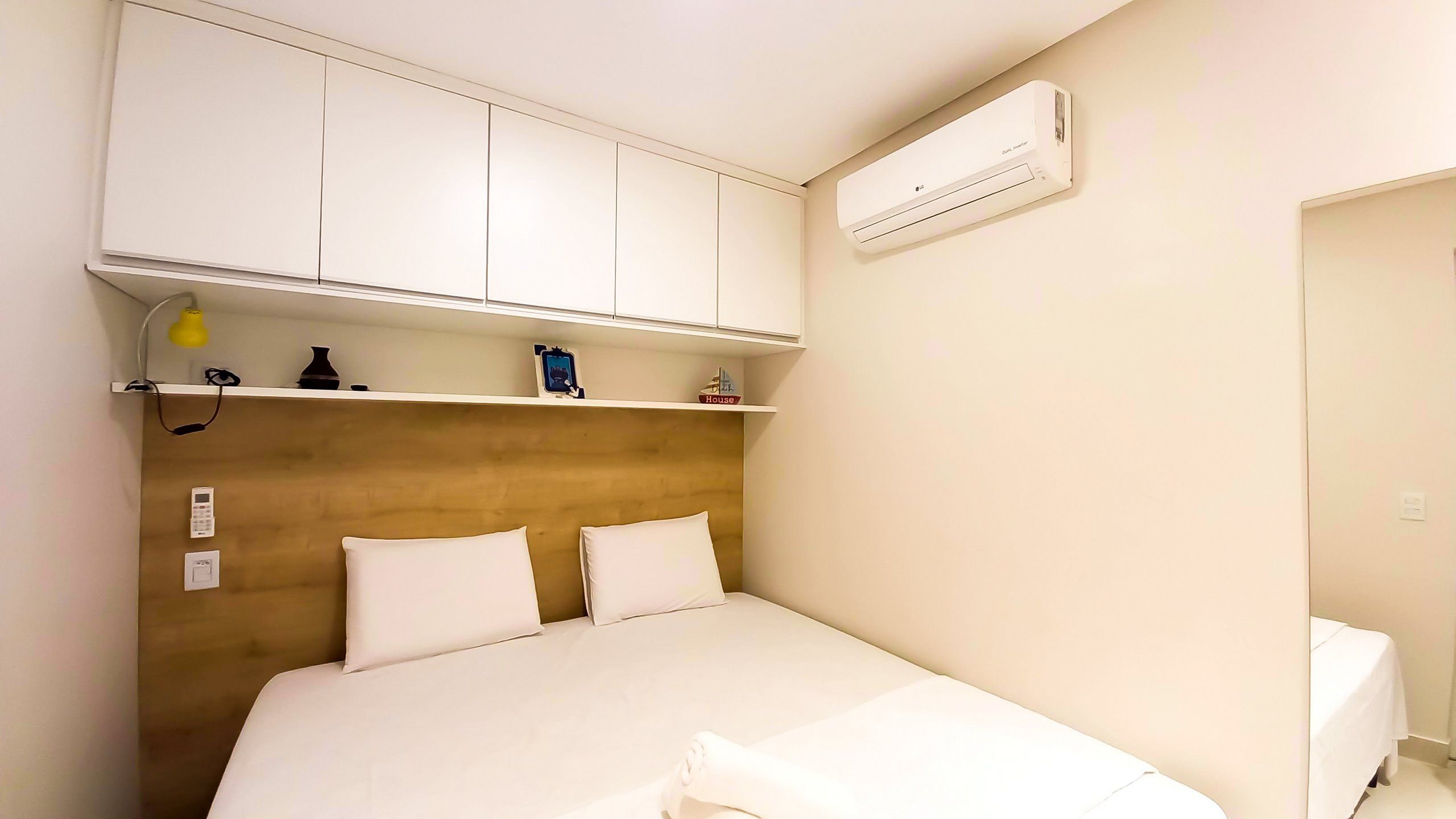 A hotel room with a bed, white headboard, bedside tables, a shelf with items, an air conditioner, and a door.