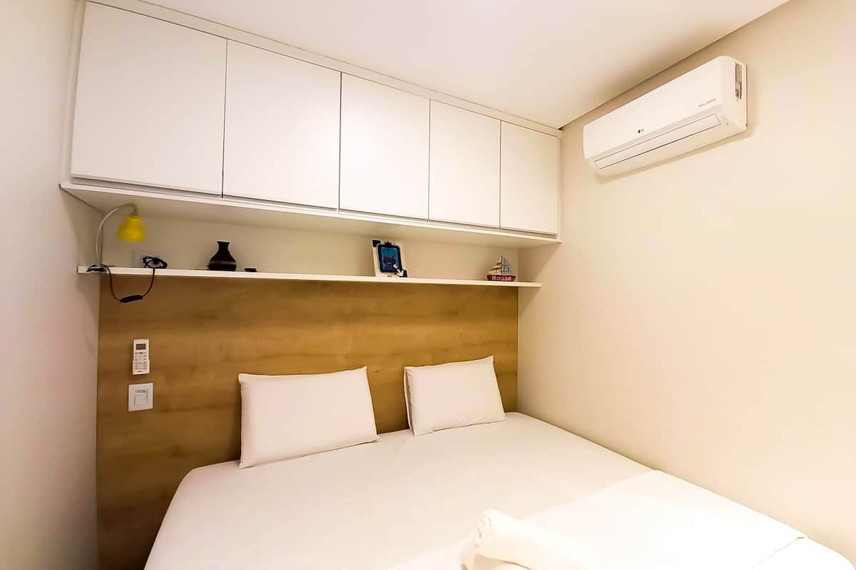 A hotel room with a bed, white headboard, bedside tables, a shelf with items, an air conditioner, and a door.