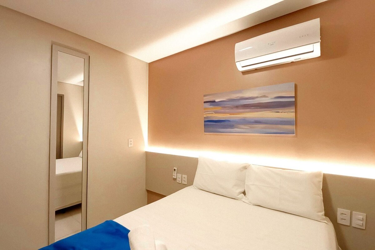A hotel room with a bed, an air conditioning unit, a painting on the wall, and a door leading to another room.