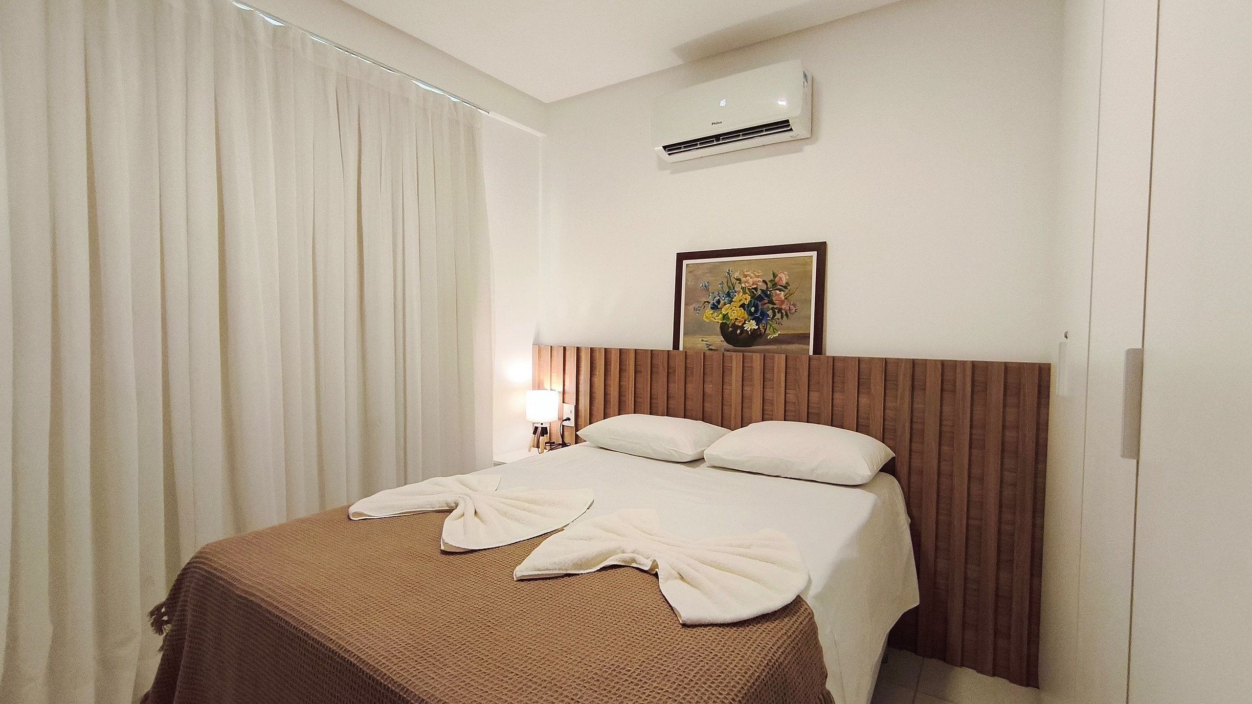 A hotel room with a bed, curtains, an air conditioner, a nightstand, a lamp, and a framed picture on the wall.