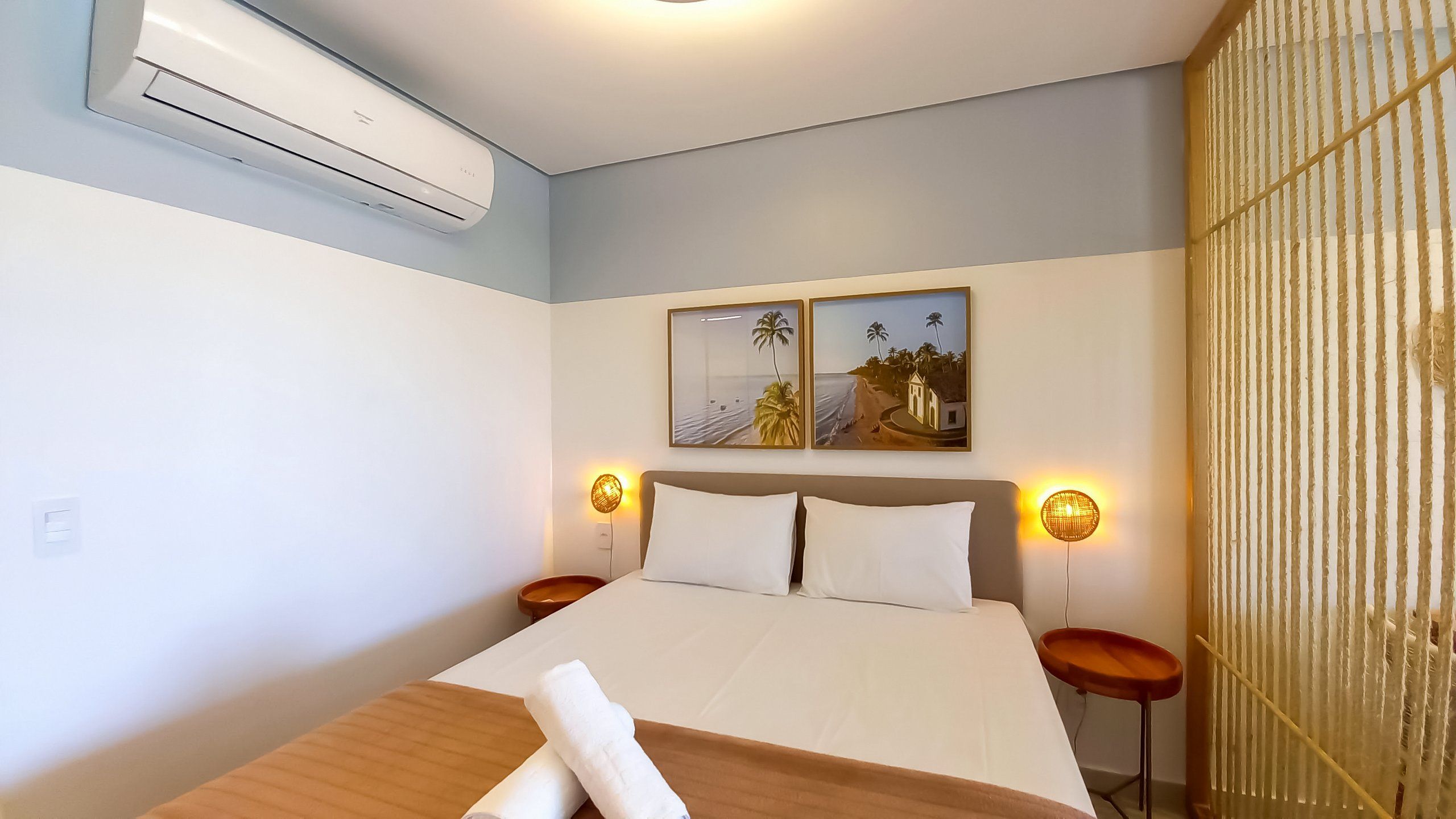 A hotel room with a bed, two bedside tables, an air conditioning unit, and two wall-mounted lamps.