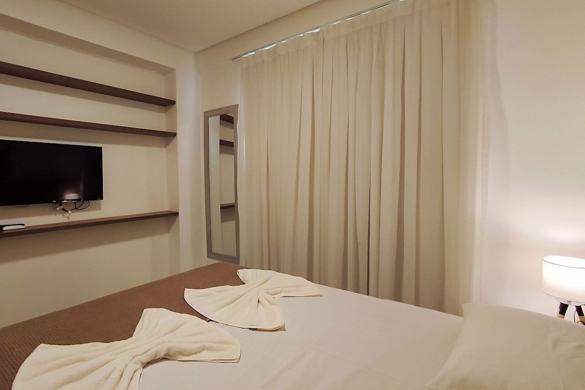 A hotel room with a bed, a TV mounted on the wall, a mirror, and a window with curtains.