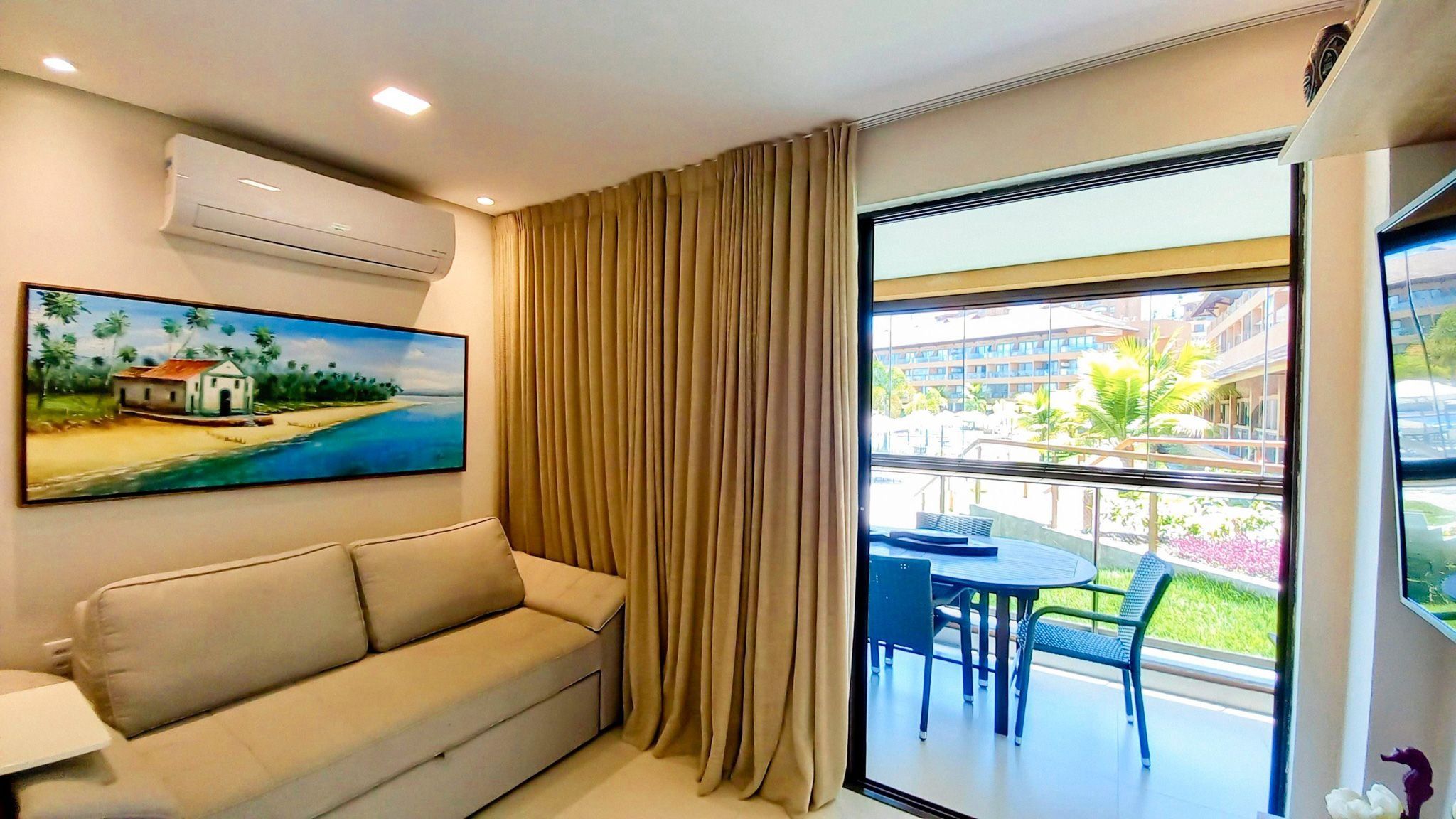 A hotel room with a beige sofa, a painting of a beach scene, a large window with curtains, and a balcony with outdoor furniture.