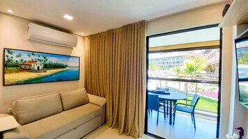 Habitación de hotel con sofá color beige, un cuadro de una playa, un ventanal con cortinas y un balcón con mobiliario exterior.